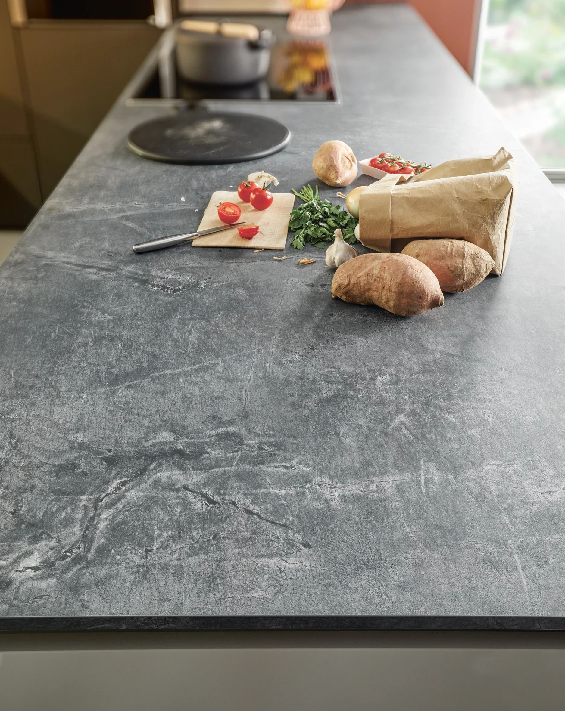 Schüller: Arbeitsplatte in dunkler Steinoptik im Fokus, darauf Gemüse und Brot, Kochfeld im Hintergrund